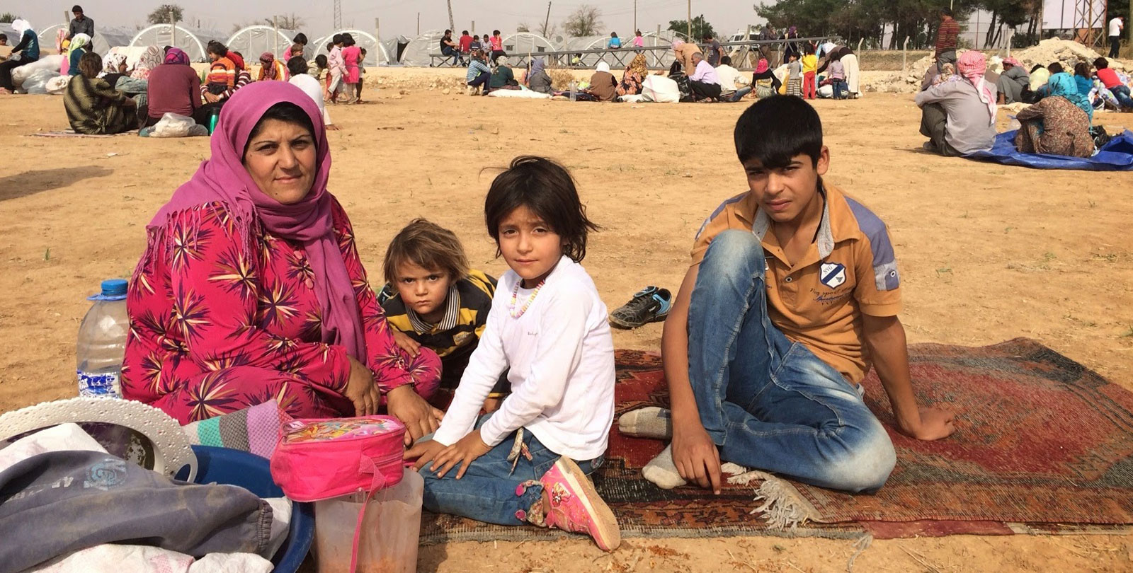 Une mère syrienne est assise avec ses enfants et quelques biens dans un camp de réfugiés sur le sol à l'air libre.