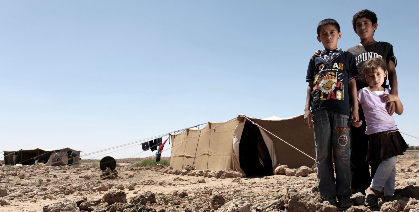 Trois enfants sont debouts devant une tente dans un camp de réfugiés.